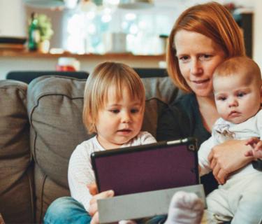 Woman sitting on a couch holding a baby, a toddler, and a tablet
