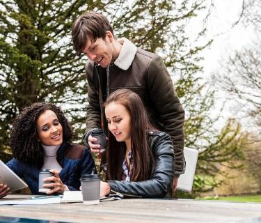 3 students sitting at a park table looking at notes and drinking coffee