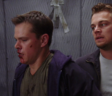 Two men stand in front of grey wall. One man has a cut chin