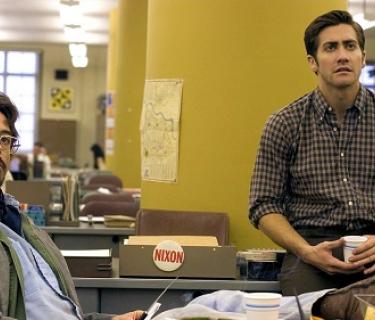 Two men sitting in an office, one in a chair and one on a desk