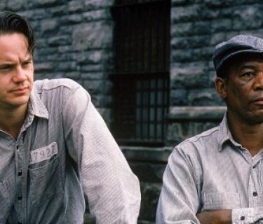 Two men in prison suits standing against a grey brick wall