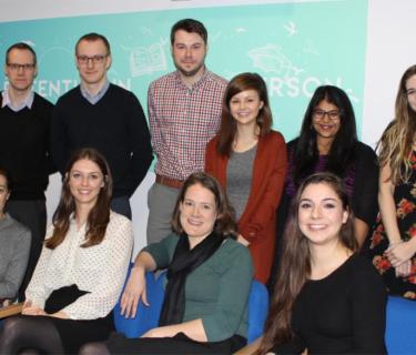 Group of 12 people posing for a group photograph. 4 people are sitting in blue chairs and 8 people are standing behind