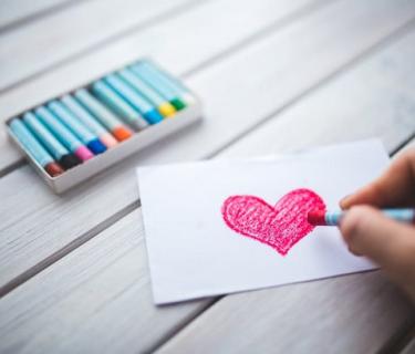 Person colouring a heart on a piece of white paper with box of crayons nearby