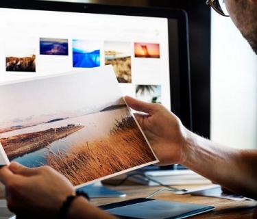 Man holding a printed image with more images on a computer montior in the background