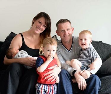 Family photo of a mother, father, infant, and two young boys all sitting on a grey couch