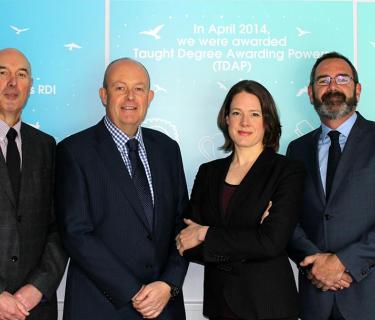 Four people in suits standing in front of Arden University posters