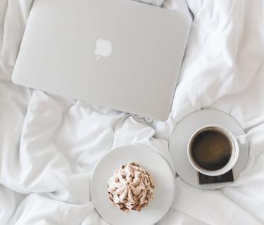 Looking down at an apple laptop, coffee on a saucer, and pastry on a plate, all on top of a bed